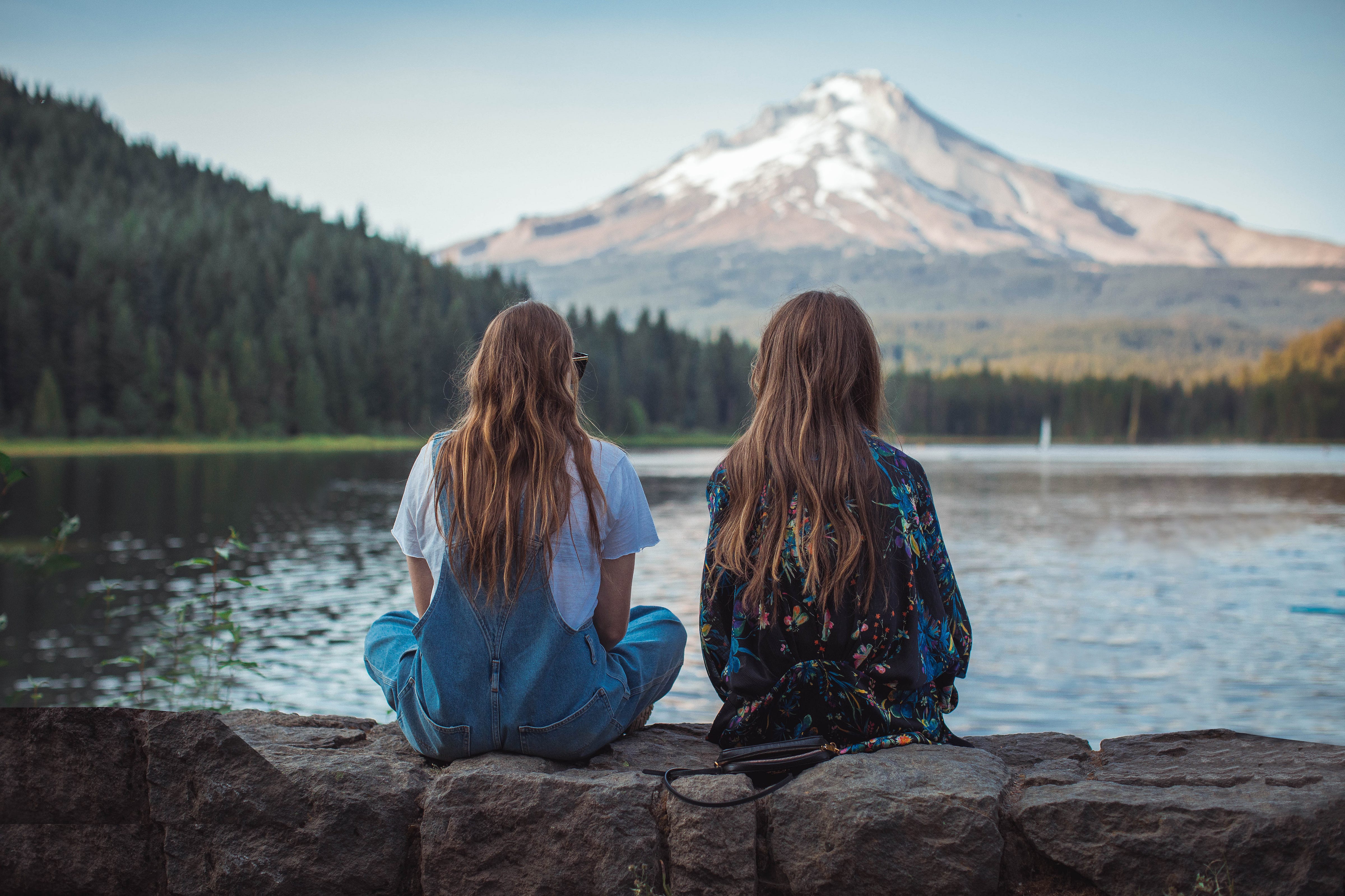 Two girls sitting on a stone wall in the country, looking across a lake at a snow capped mountain.